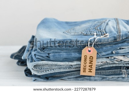 Stack of blue jeans and tag with inscription second hand on white background. Second hand clothing shop. Circular fashion, eco friendly sustainable shopping, thrift stores concept.
