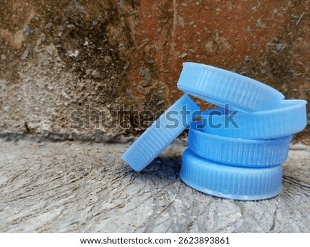 Stack of blue plastic bottle caps on cement floor near a rough, mossy wall.
