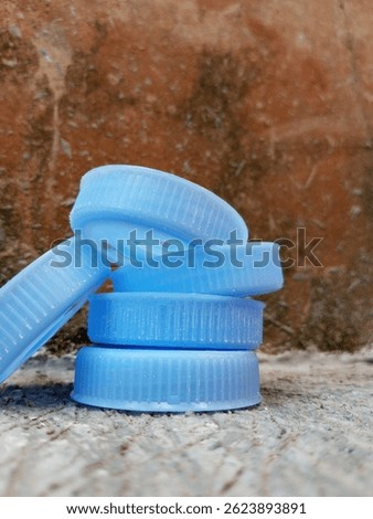 Stack of blue plastic bottle caps on cement floor near a rough, mossy wall.
