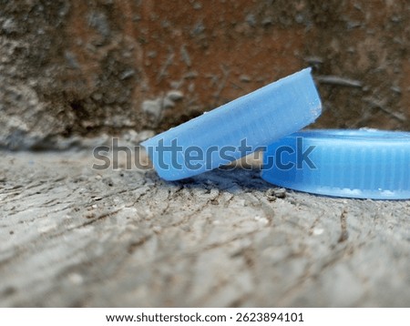 Stack of blue plastic bottle caps on cement floor near a rough, mossy wall.
