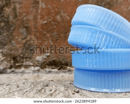 Stack of blue plastic bottle caps on cement floor near a rough, mossy wall.
