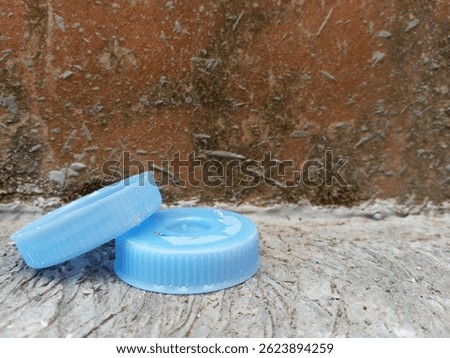 Stack of blue plastic bottle caps on cement floor near a rough, mossy wall.
