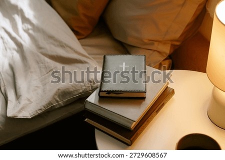 Stack of books with closed Bible featuring cross symbol resting on top, placed on round bedside table next to illuminated lamp and unmade bed with pillows and blanket visible 