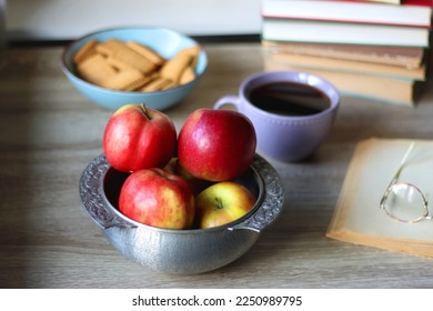 Stack of books, e-reader, reading glasses, bowl of biscuits, apples and cup of tea on the table. Bookshelf in the background. Selective focus.
