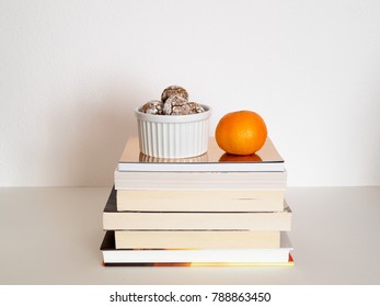 A stack of books in front of a white background with a tangerine and a cup of biscuits on it.