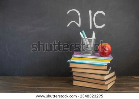 Stack of books and notebooks, red apple, pencil holder on wooden desk in front of blackboard with Hebrew letters 