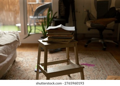 Stack of books on stool, soft morning light through glass door, patterned rug, scattered cushions, open book on top, houseplant blurred in background, calm slow living atmosphere, warm