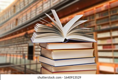 Stack Of Books With An Opened Book In Front Of Shelves Of A Large Library