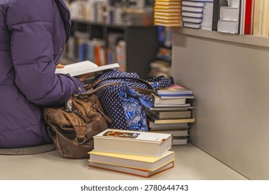 Stack of books and reader sitting in bookstore, surrounded by bags and novels. Reading moment, peaceful bookstore atmosphere, love for books, reading fiction concept. Real scene in store