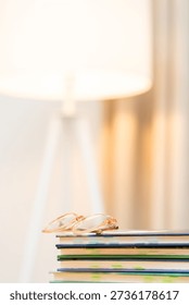 A stack of books sits on a table with glasses resting on top. A lamp lights up the area, creating a warm space for reading. This setting feels inviting and perfect for a reader.