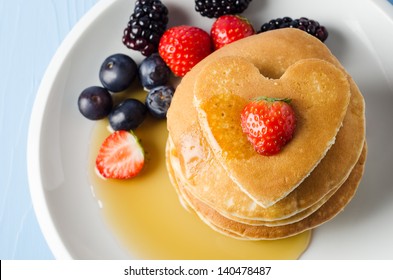A Stack Of Breakfast Pancakes Topped With A Strawberry, On A White Plate With Summer Fruits.  Maple Syrup Drizzles Down Over The Pancakes From Above.  Pale Blue Wooden Table And Background.