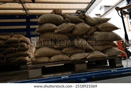 stack of burlap sandbags sits on a wooden pallet inside a truck, ready for transport. A worker is seen using a pallet jack to move the sandbags, likely for flood protection or emergency use