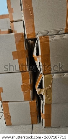 Stack of cardboard boxes with damaged and crushed corners, sealed with brown packing tape. Captured in a warehouse or storage setting, highlighting packaging issues or logistics mishandling.