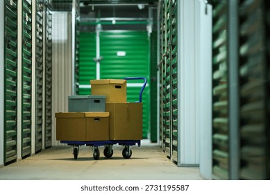 Stack of cardboard boxes on blue hand truck standing in corridor of self storage facility, green metal storage unit doors lining both sides, concrete floor visible, no people present