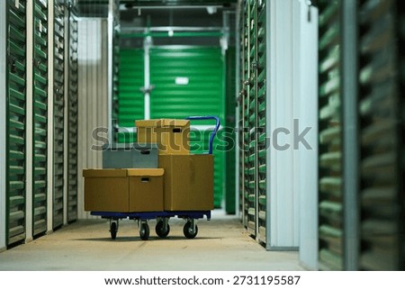 Stack of cardboard boxes on blue hand truck standing in corridor of self storage facility, green metal storage unit doors lining both sides, concrete floor visible, no people present