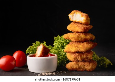 A stack of chicken nuggets and a bowl with ketchup and lettuce on a dark background.