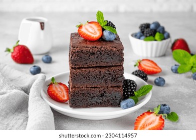 A stack of chocolate brownies with chocolate glaze, fresh berries and mint leaves on top on a white plate on a gray concrete background. Delicious dessert