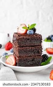 A stack of chocolate brownies with chocolate glaze, fresh berries and mint leaves on top on a white plate on a gray concrete background. Delicious dessert