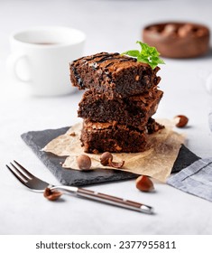 A stack of chocolate brownies with mint, hazelnuts and salted caramel on baking paper on a light background with a cup of coffee. The concept of delicious and sweet homemade baked goods for breakfast.