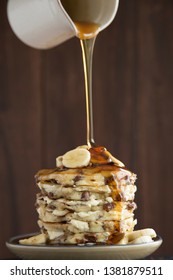 A Stack Of Chocolate Chip Banana Pancakes With A Pitcher Pouring Maple Syrup