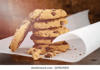 Stack of chocolate chip cookies on white curling baking paper against a dark wood background, with applied vintage style filters and added light stream. 