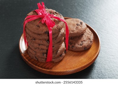 Stack of chocolate chip cookies wrapped in a red ribbon on wooden plate on black stone background