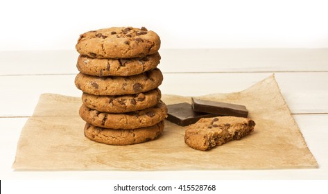A stack of chocolate chips cookies and a half of one, with a couple of pieces of dark chocolate, on a sheet of baking paper, with copyspace
