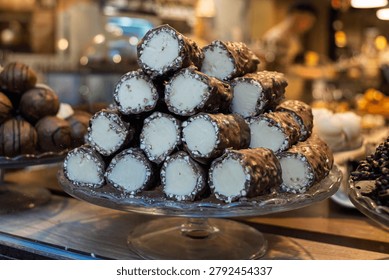 Stack of chocolate-covered pastry rolls filled with cream on a glass cake stand in a bakery or candy shop