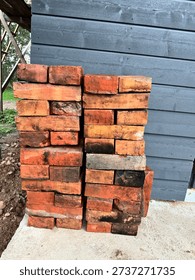Stack of clay bricks placed on a concrete surface beside a building wall. The bricks are arranged in two vertical piles, positioned next to exterior siding made of horizontal wooden boards. The units vary in tone due to firing marks, soot traces, and previous use. Some pieces show signs of mortar residue, indicating they may have been removed from an earlier structure. The stack stands near ground soil and construction material, suggesting ongoing building or renovation work.

Each brick is rectangular and uniform in size, designed for masonry applications such as walls, foundations, fireplaces, or paving. The arrangement demonstrates storage before installation, with the materials kept close to the work area. The scene reflects a construction site environment, where supplies are organized for easy access during masonry tasks. The concrete slab provides a stable base, preventing contact with soil moisture.

The background shows part of a residential structure with siding panels and a small section of roof or framework visible to the side. No workers are present in the frame, placing focus on the building materials themselves. The composition highlights texture, stacking method, and the practical aspects of construction logistics.

This image can illustrate themes such as home improvement, renovation projects, masonry work, building supplies, property maintenance, and material reuse. It is suitable for editorial content related to construction industry topics, contractor services, architectural planning, or do-it-yourself projects. The clear presentation of the stacked bricks allows space for text placement and design elements, making the photograph adaptable for marketing materials, informational articles, or trade publications connected to housing development and structural repair.