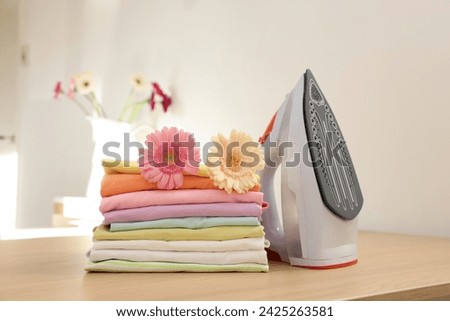 Stack of clean clothes, modern iron and flowers on wooden table