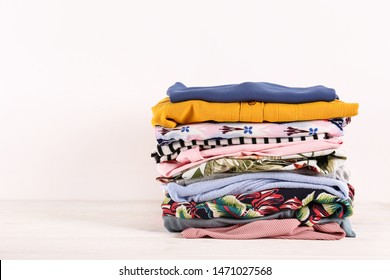 Stack Of Clean Freshly Laundered, Neatly Folded Women's Clothes On Wooden Table. Pile Of Shirts, Dresses And Sweaters On White Board, Concrete Wall Background. Copy Space, Close Up, Top View.