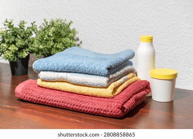 Stack of colorful folded towels with detergent containers placed on wooden table near potted plant
