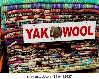 stack of colorful, intricately patterned woolen fabrics made from yak wool on display. A sign labeled Yak Wool with an image of a yak prominently identifies material, showcasing natural, cozy textures - Powered by Shutterstock