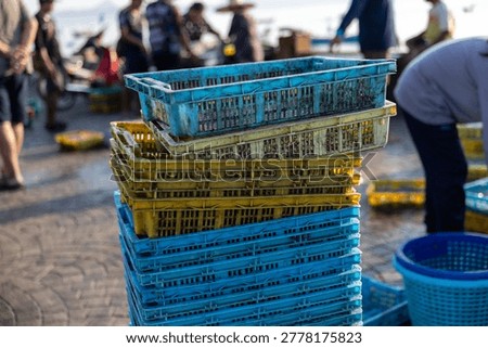 Stack of colorful plastic crates at bustling fishing port with people working in background creating lively energetic atmosphere under bright morning sunlight near ocean