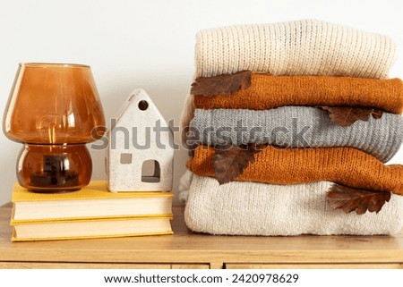 Stack of cozy knitted warm sweater with fall leaf, books, decorative broun lamp and ceramic house. Cozy hygge concept. Home autumn composition in the interior of the room. Simple minimal home cozy.