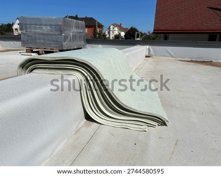 A stack of cut protective material sheets folded on the parapet of a flat membrane roof. These pads are used to be placed under concrete ballast blocks to prevent damage to the roof's waterproofing layer during solar panel installation
