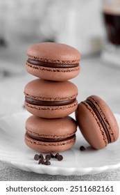 A stack of dark chocolate macarons with chocolate ganache filling on a white plate on a gray background. French meringue cookies macaron. Copy space