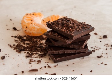 Stack of dark chocolate pieces on a light parchment paper with slices of mandarin in the background and chocolate chips