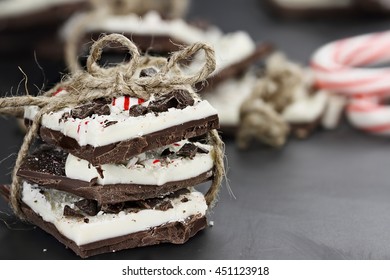 Stack of dark and white chocolate peppermint bark tied together with twine over a black background with more peppermint candies in the background. Extreme shallow depth of field.