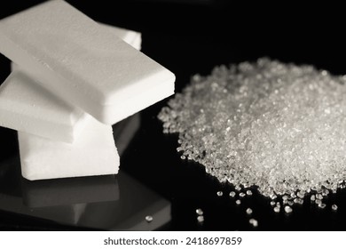 A stack of dextrose glucose tablets and a pile of white granulated sugar on black glass.