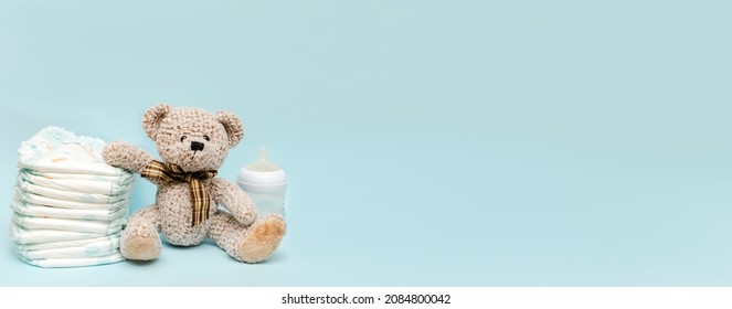 Stack of diapers with cute teddy bear toy, bottle of milk on table. set for infant boy girl for baby shower present gift on blue background, banner, copy space. Healthcare medical, hygiene concept.
