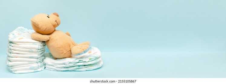 Stack of diapers with cute teddy bear toy lying on table. set for infant newborn boy girl for baby shower present gift on blue background with copy space. Healthcare medical, hygiene concept.