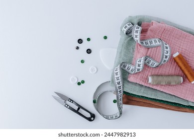 a stack of different fabric scraps lies on a white background, on top are two skeins of thread, tailor scissors and a measuring tape. buttons nearby. shot from above