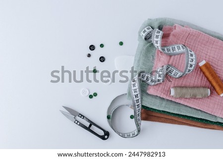 a stack of different fabric scraps lies on a white background, on top are two skeins of thread, tailor scissors and a measuring tape. buttons nearby. shot from above