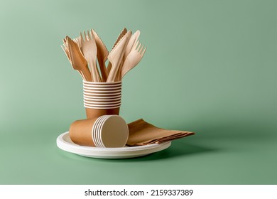 Stack of eco-friendly disposable tableware. Wooden forks and knives, paper cups and plates against green background. Biodegradable cutlery and dishes for picnics, takeaways. Copy space. Front view.