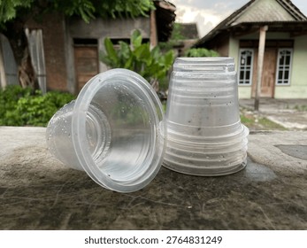 Stack of empty clear disposable plastic cups on a stone ledge outdoors with a blurred village house and greenery in the background.