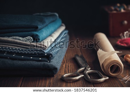 Stack of fabrics for sewing  and sewing items - tailoring scissors, including pins, cutting knife, paper for sewing pattern. Wooden box of spools of thread on background.