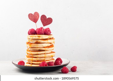 Stack of fluffy pancakes with syrop, decorated with red hearts and raspberries. Homemade present for Saint Valentine day. Light background. Copy space.