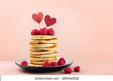 Stack of fluffy pancakes with syrup, decorated with red glitter pepper hearts and raspberries. Homemade present for Saint Valentine day. Light peach colour background. Copy space.