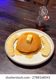 Stack of fluffy pancakes topped with fresh banana slices, melting butter, and drizzled with maple syrup on a rustic wooden table.